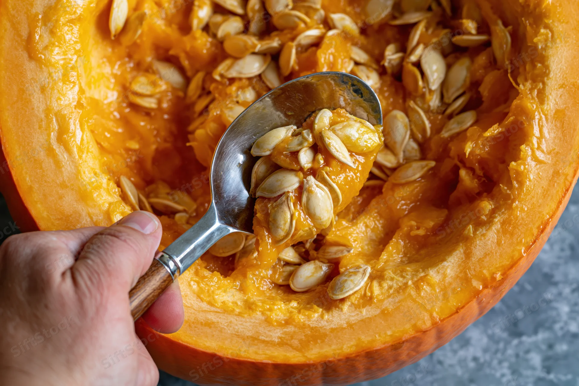 Hand scooping wet pumpkin seeds and pulp from a halved pumpkin on a countertop