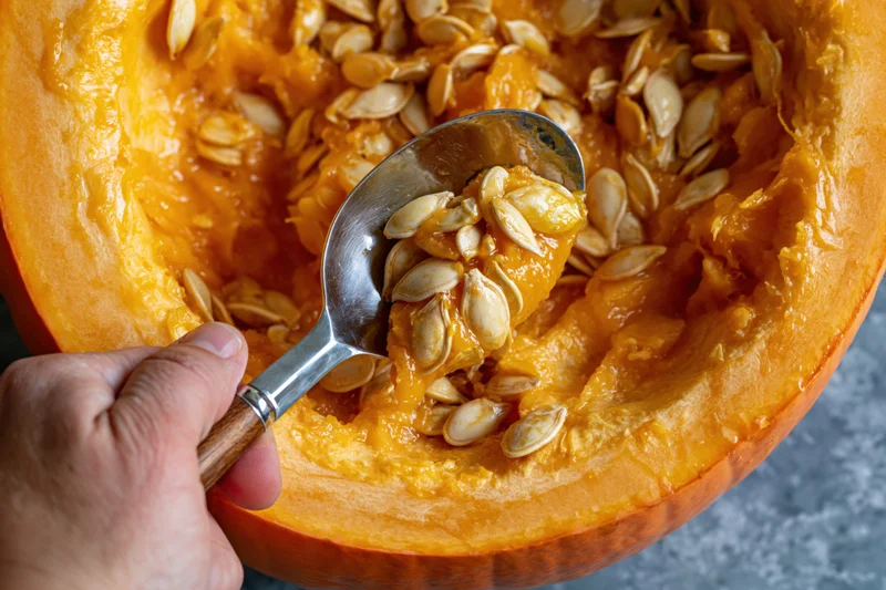 Hand scooping wet pumpkin seeds and pulp from a halved pumpkin on a countertop