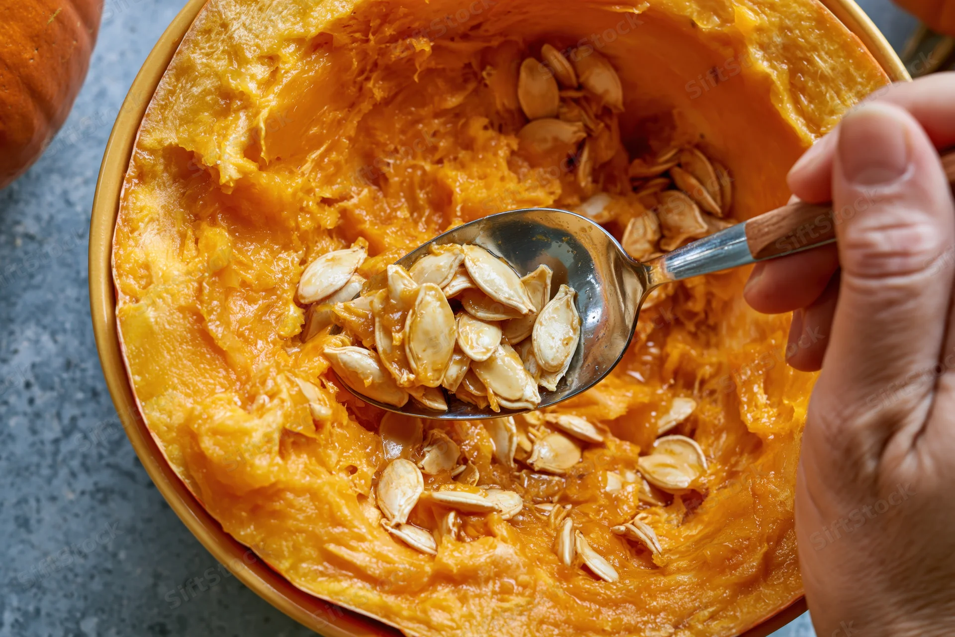 Hand scooping wet pumpkin seeds from a hollowed pumpkin with a metal spoon on a gray surface