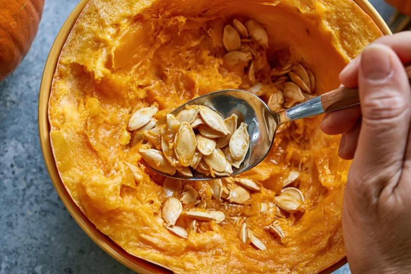 Hand scooping wet pumpkin seeds from a hollowed pumpkin with a metal spoon on a gray surface