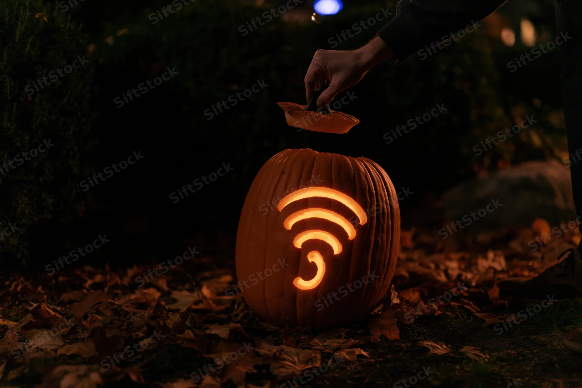 Hand lifting stem lid off glowing jack-o-lantern carved with wifi symbol in garden at night