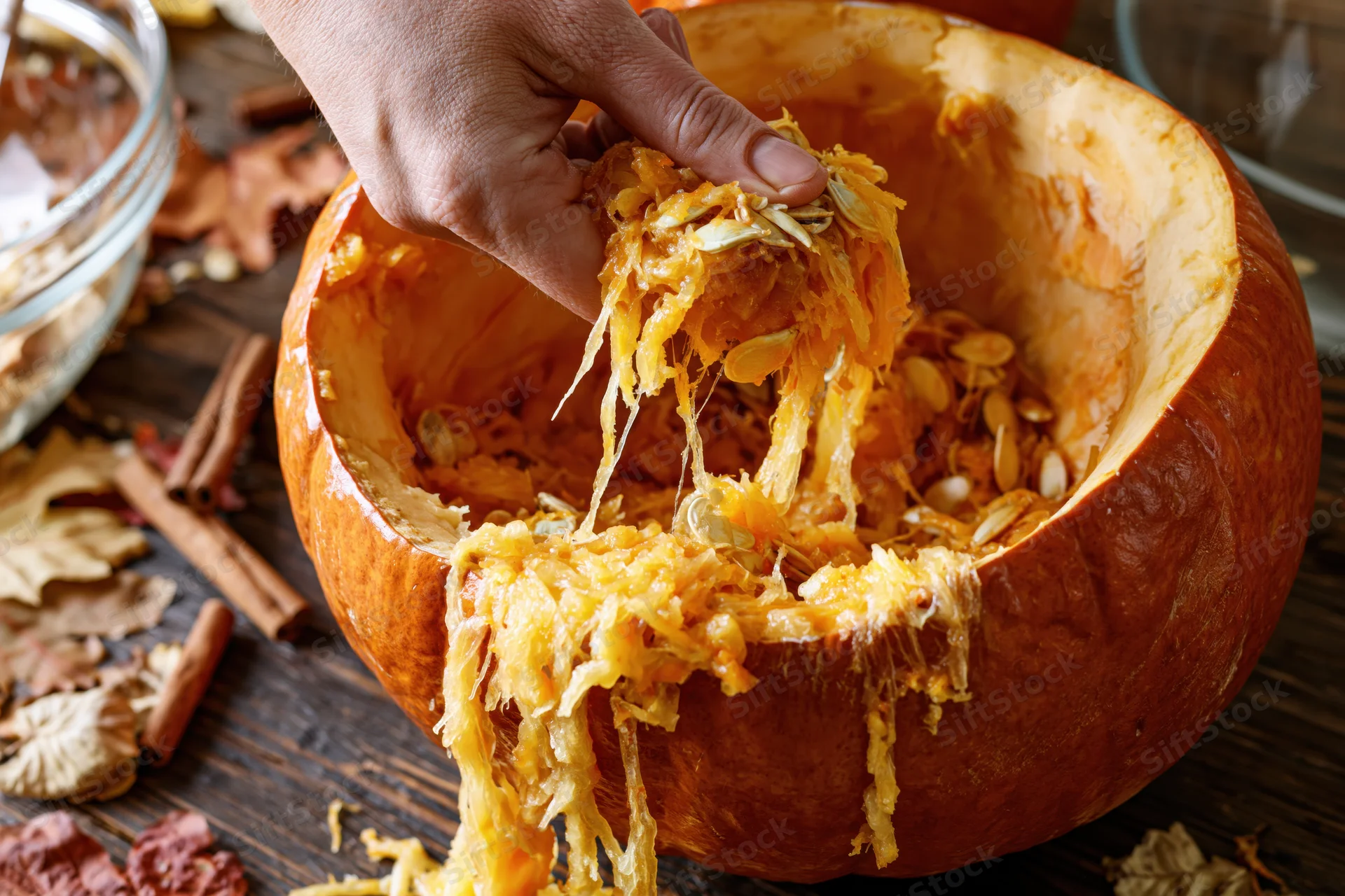 Hand pulling fibrous orange pumpkin guts and seeds from hollowed pumpkin on wooden table with autumn leaves and cinnamon sticks