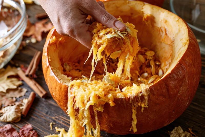 Hand pulling fibrous orange pumpkin guts and seeds from hollowed pumpkin on wooden table with autumn leaves and cinnamon sticks