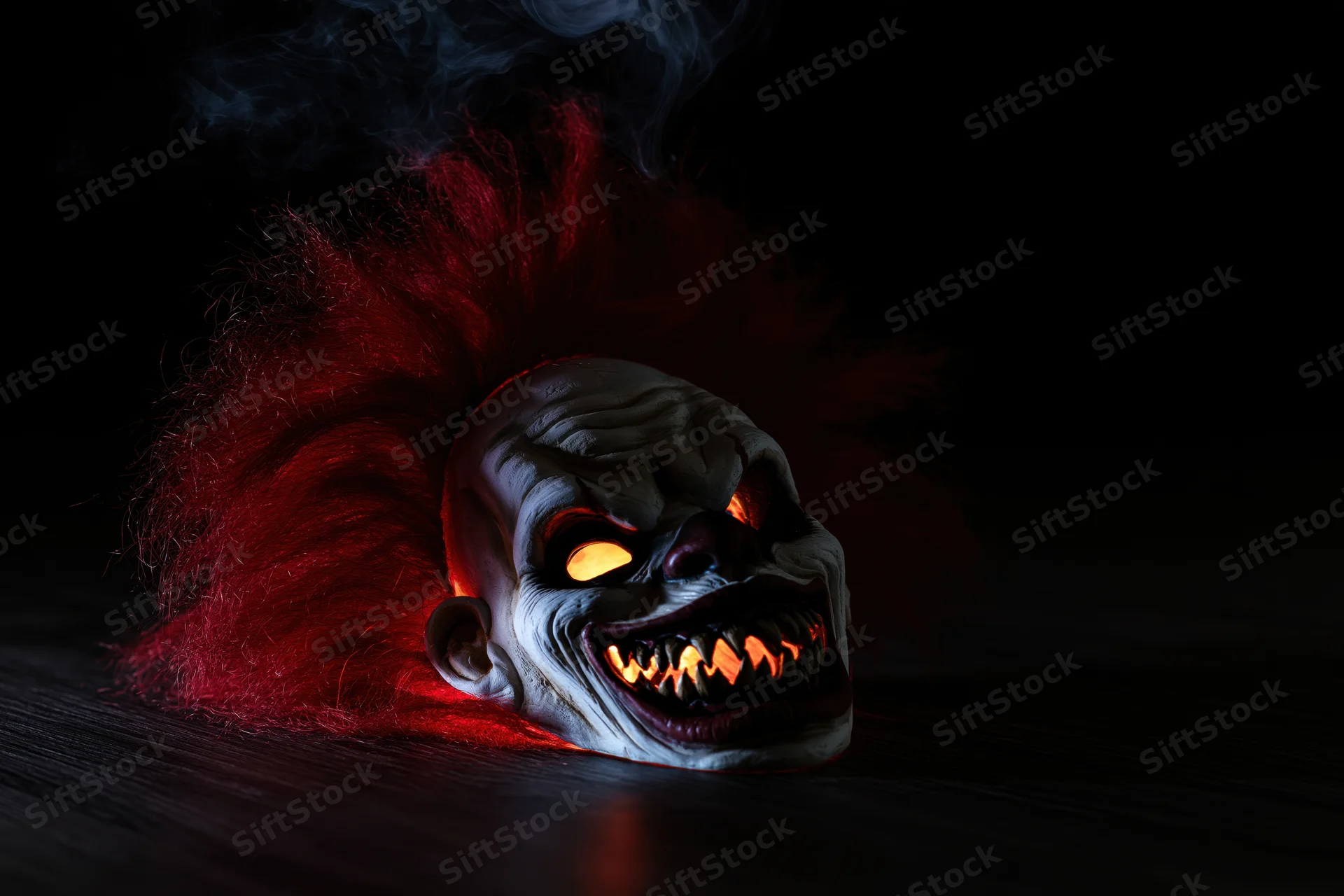 Horror clown mask with glowing eyes and red frizzy hair resting on dark wooden surface