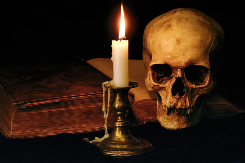 Human skull beside an open aged book with a lit candle on a dark surface
