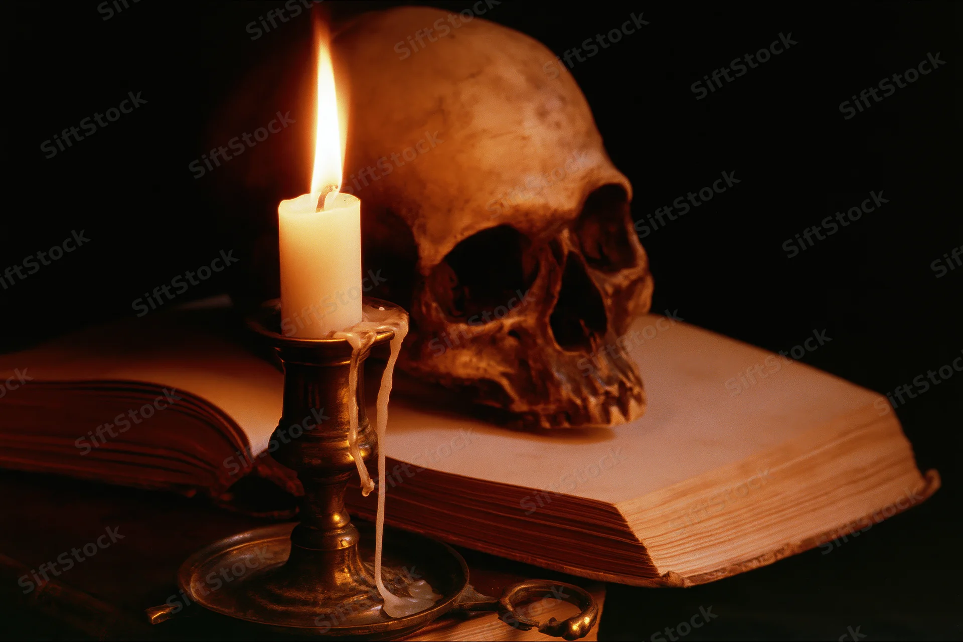 Human skull resting behind an open aged book with a lit candle on a brass candlestick