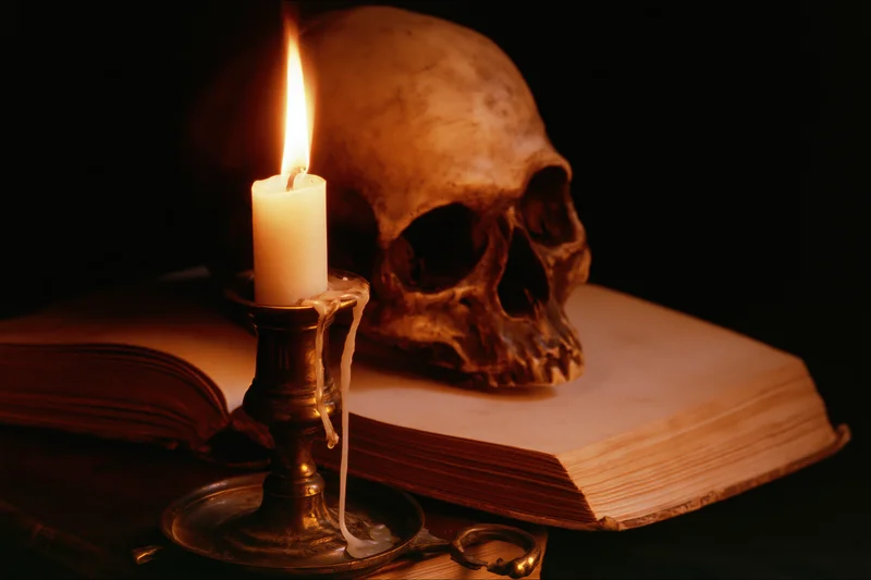 Human skull resting behind an open aged book with a lit candle on a brass candlestick