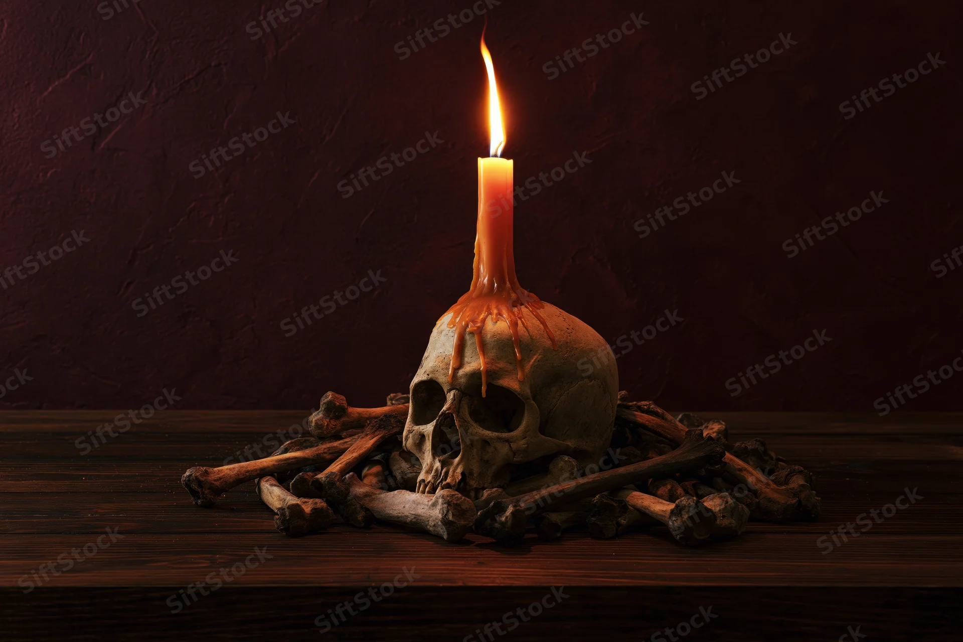 Human skull with lit orange candle melting on top, surrounded by bones on wooden table against dark textured wall