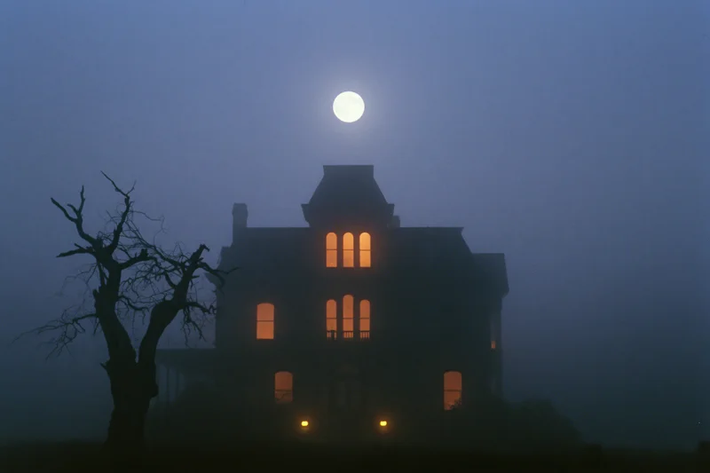 Victorian mansion silhouetted against full moon at night with fog and glowing windows
