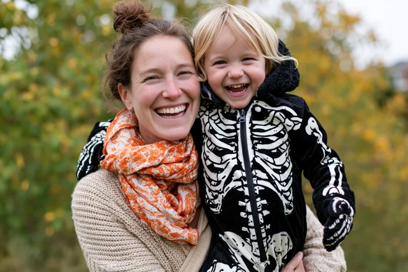 Woman laughing while holding a young child in a skeleton costume outdoors in autumn foliage