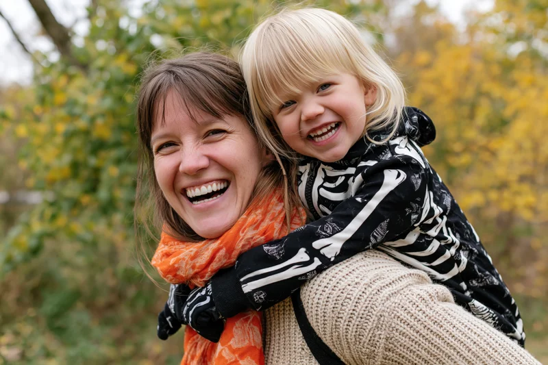 Woman carrying young child on her back, both laughing, in autumn park setting