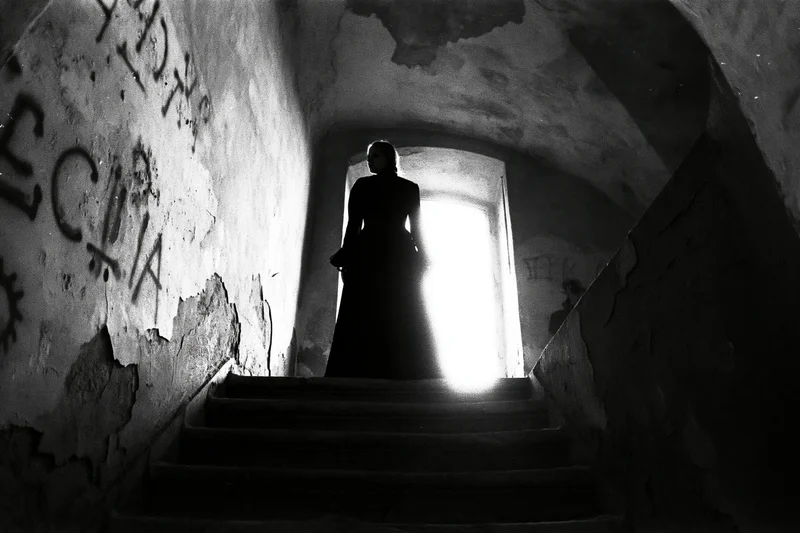Woman in silhouette standing at top of stone staircase in dimly lit abandoned tunnel with graffiti-covered walls and bright doorway ahead