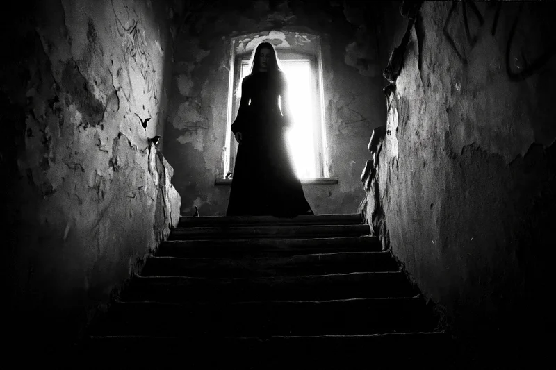 Woman in long dark dress standing at top of stairs in dimly lit abandoned hallway