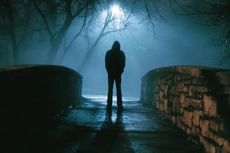 Hooded figure standing motionless on a foggy stone path at night