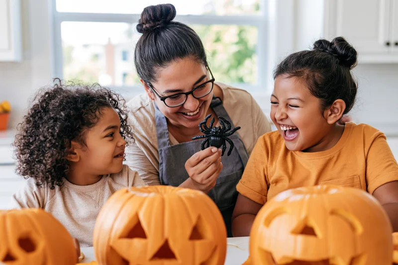 Mother holding plastic spider toward laughing daughters in kitchen with carved pumpkins