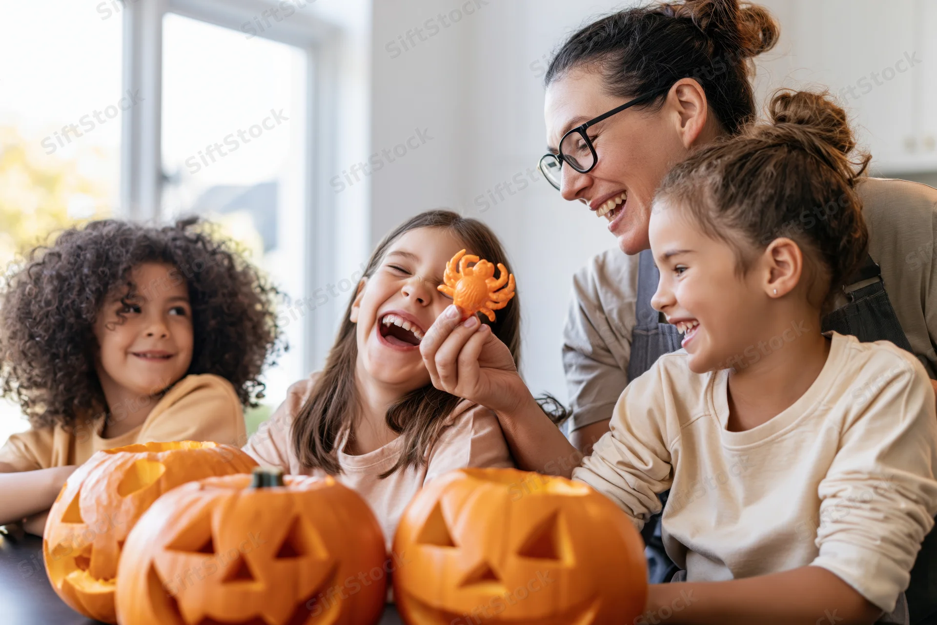 Mother holding plastic spider toward laughing daughters at table with carved pumpkins indoors