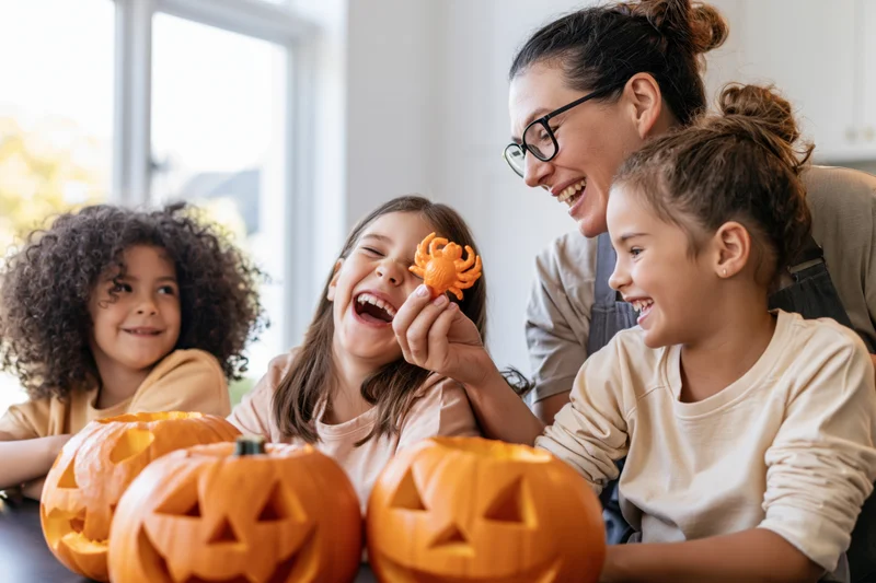 Mother holding plastic spider toward laughing daughters at table with carved pumpkins indoors