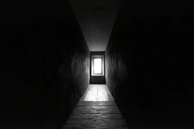 Narrow dark corridor with light at end converging toward window