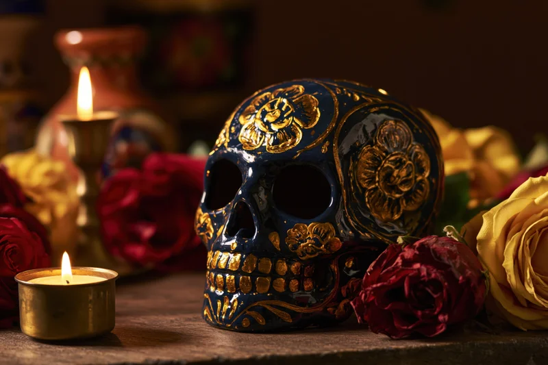 Painted ceramic sugar skull with gold floral patterns on wooden table surrounded by roses and lit candles