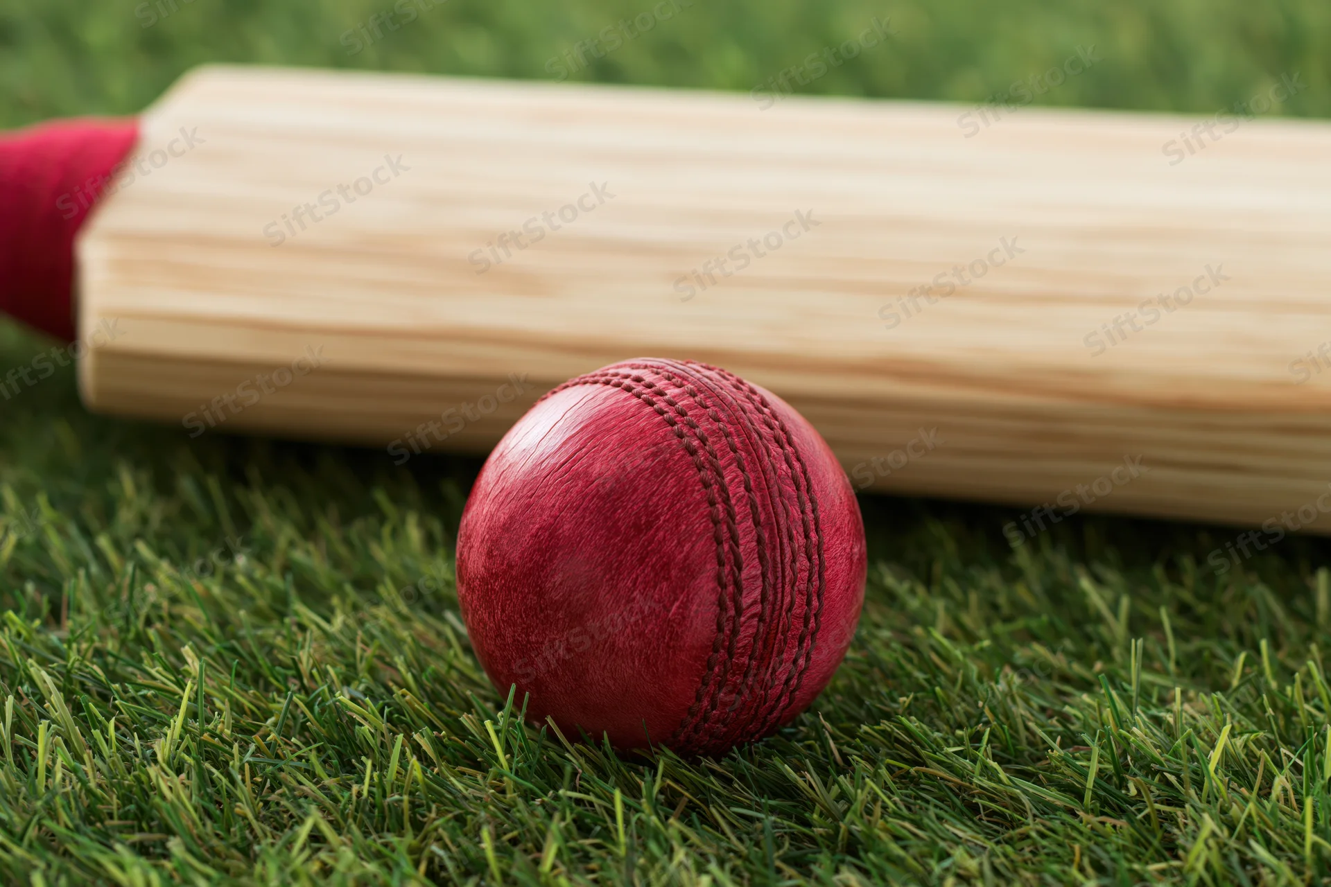 Red leather cricket ball with seam stitching resting on grass beside wooden bat