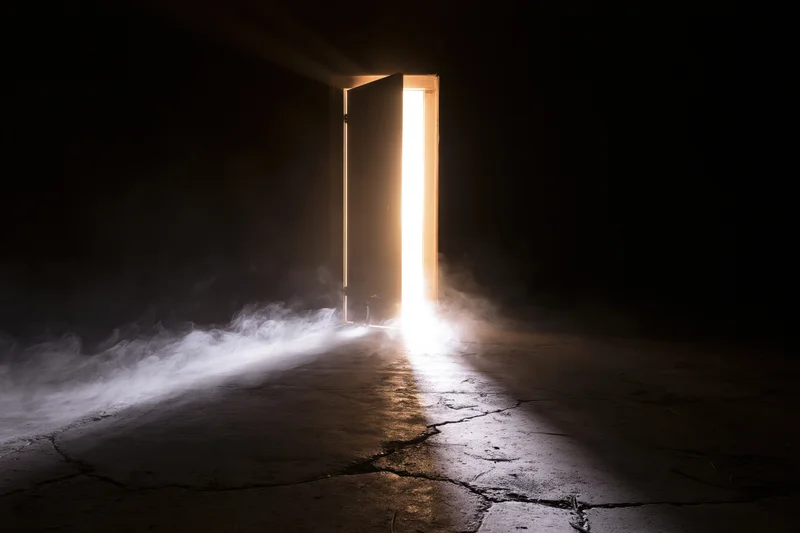 Slightly open door in dark abandoned room with light beam and smoke on cracked concrete floor