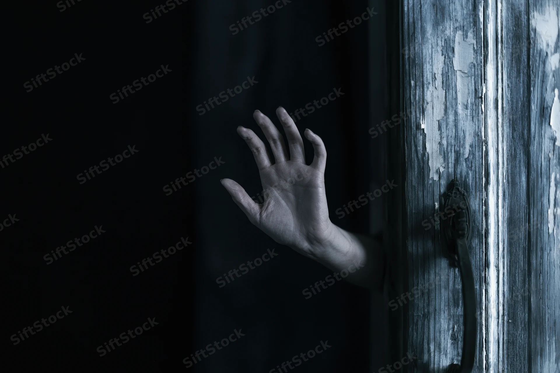 Pale hand reaching from darkness beside a weathered wooden door with peeling paint and metal handle