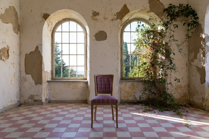 Wooden chair with faded purple fabric seat in an abandoned room with arched windows and overgrown vines