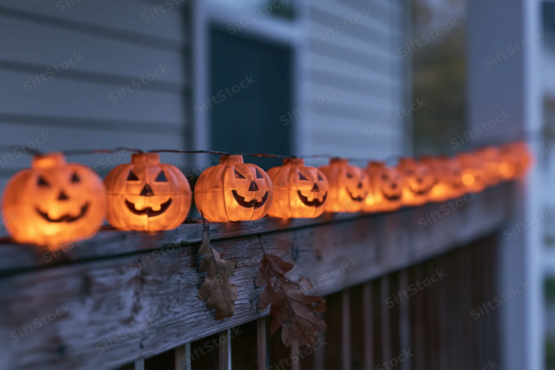 String of glowing plastic jack-o-lantern lights along wooden porch railing at twilight