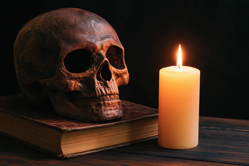 Weathered human skull resting on an old leather-bound book beside a lit candle on a dark wooden table