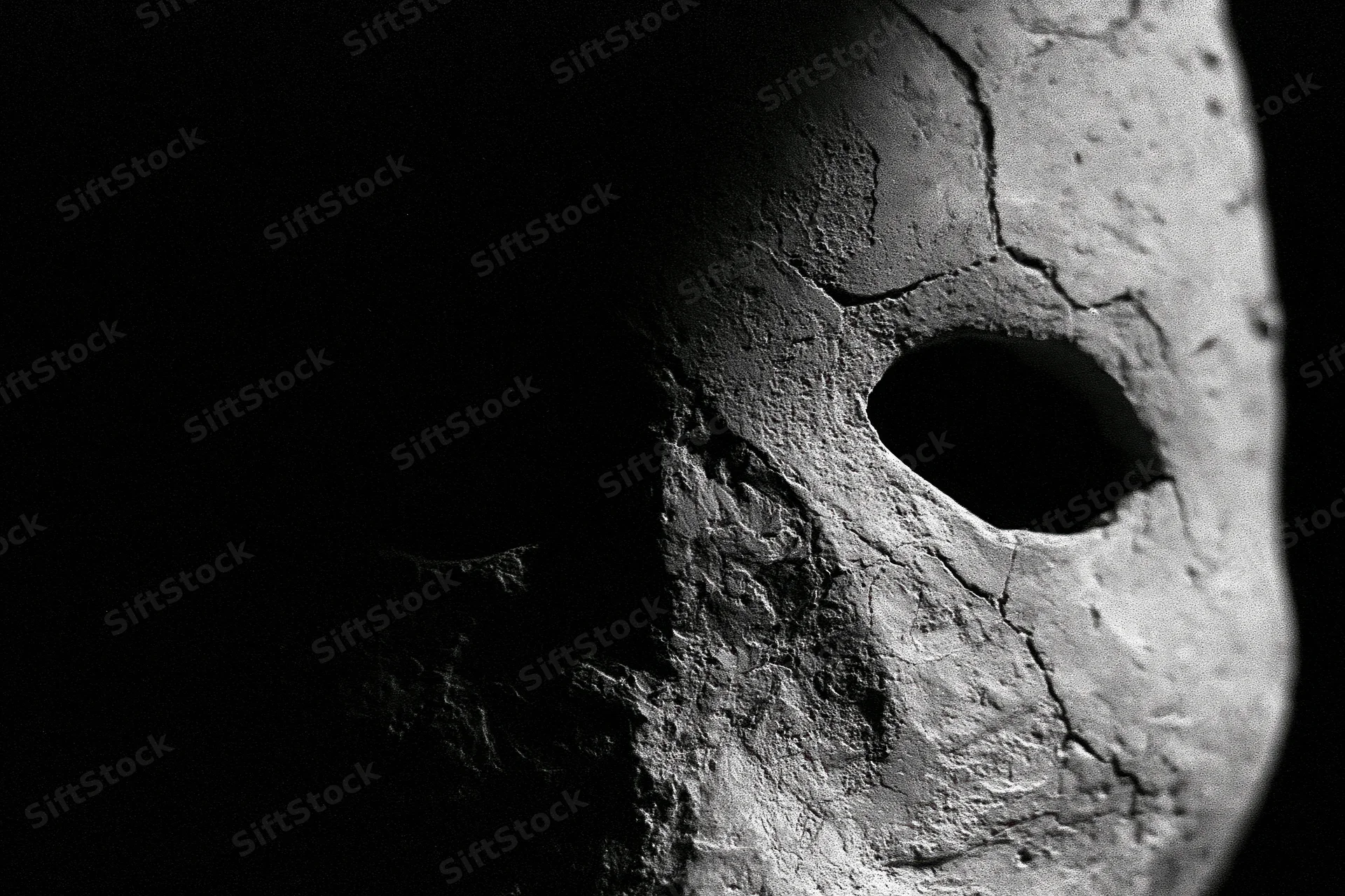 Weathered stone mask emerging from deep shadow, close-up view of cracked surface and eye opening