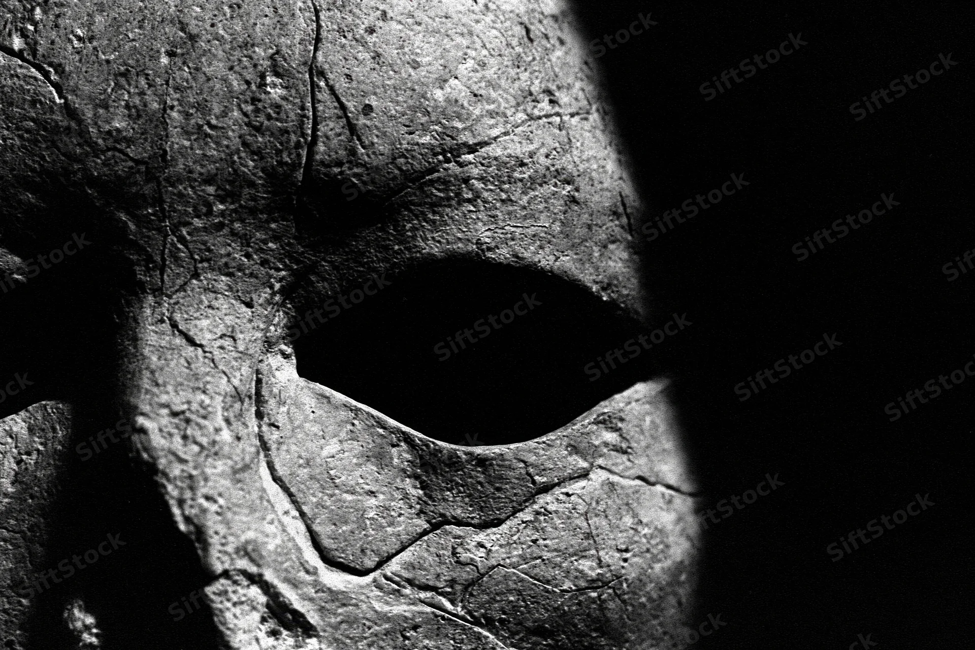 Closeup of a weathered stone mask emerging from deep shadow, showing cracked surface and eye hole