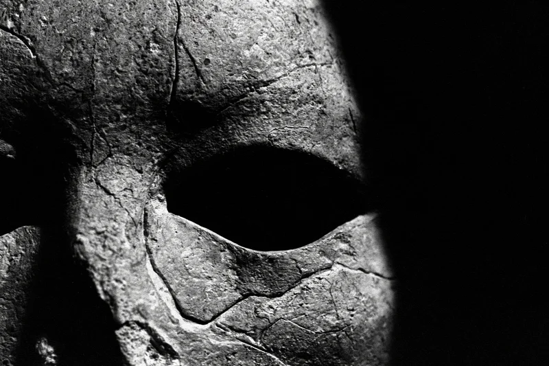 Closeup of a weathered stone mask emerging from deep shadow, showing cracked surface and eye hole
