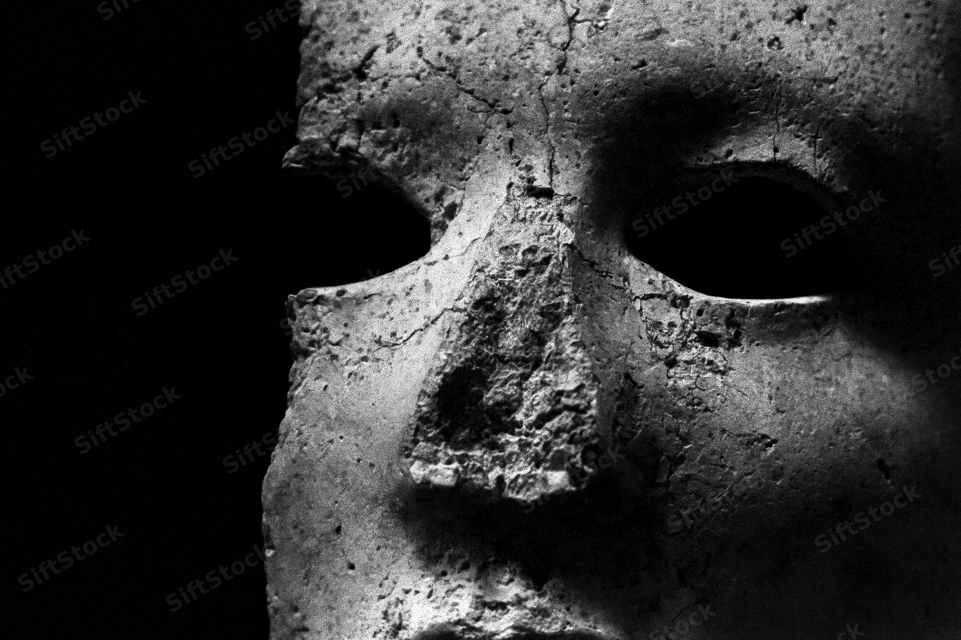 Weathered stone mask fragment emerging from deep shadow with cracked texture and hollow eyes