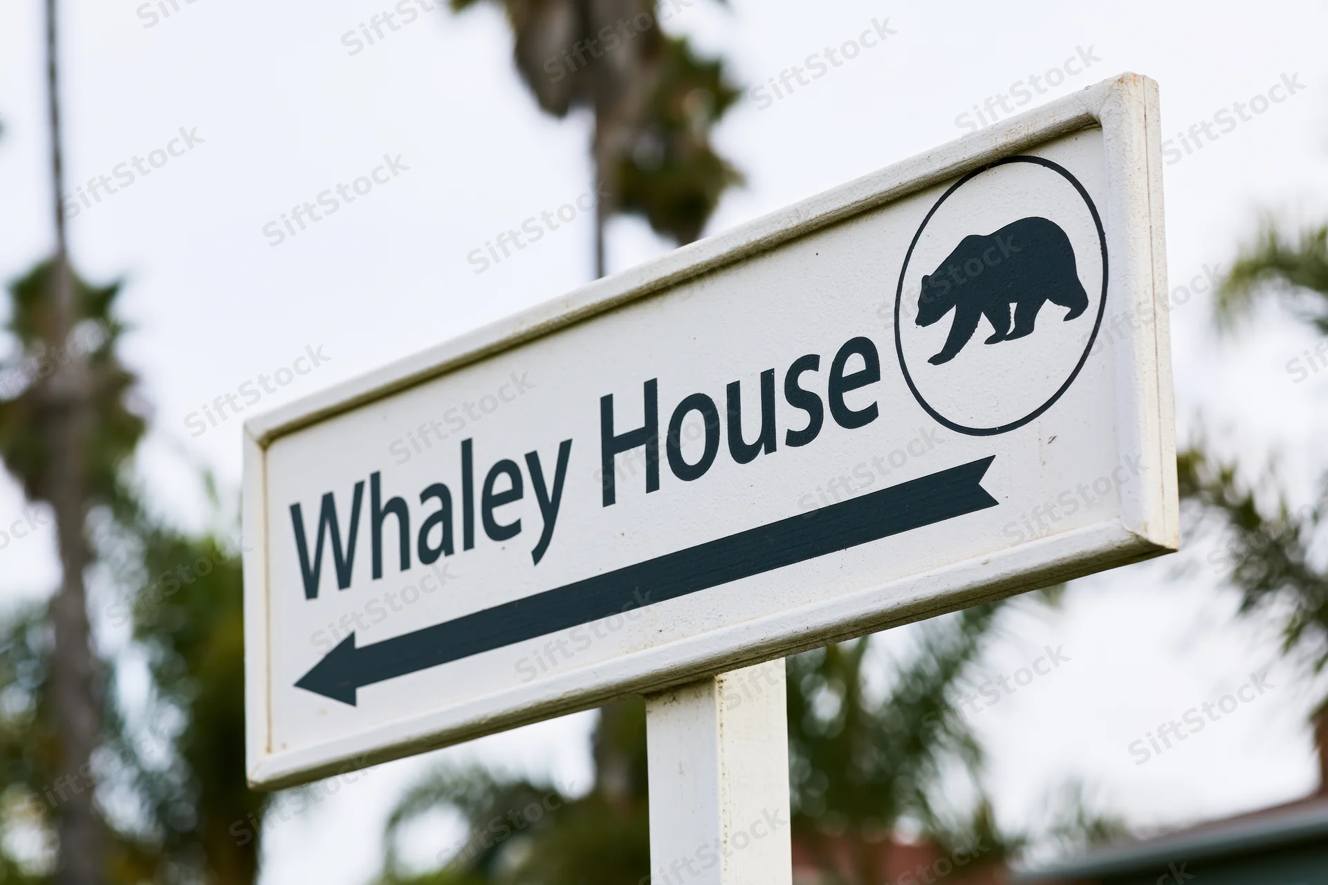 White directional sign for Whaley House with bear icon and left arrow, outdoors near palm trees