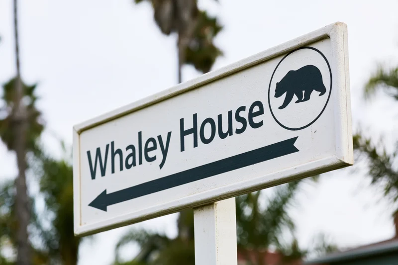 White directional sign for Whaley House with bear icon and left arrow, outdoors near palm trees