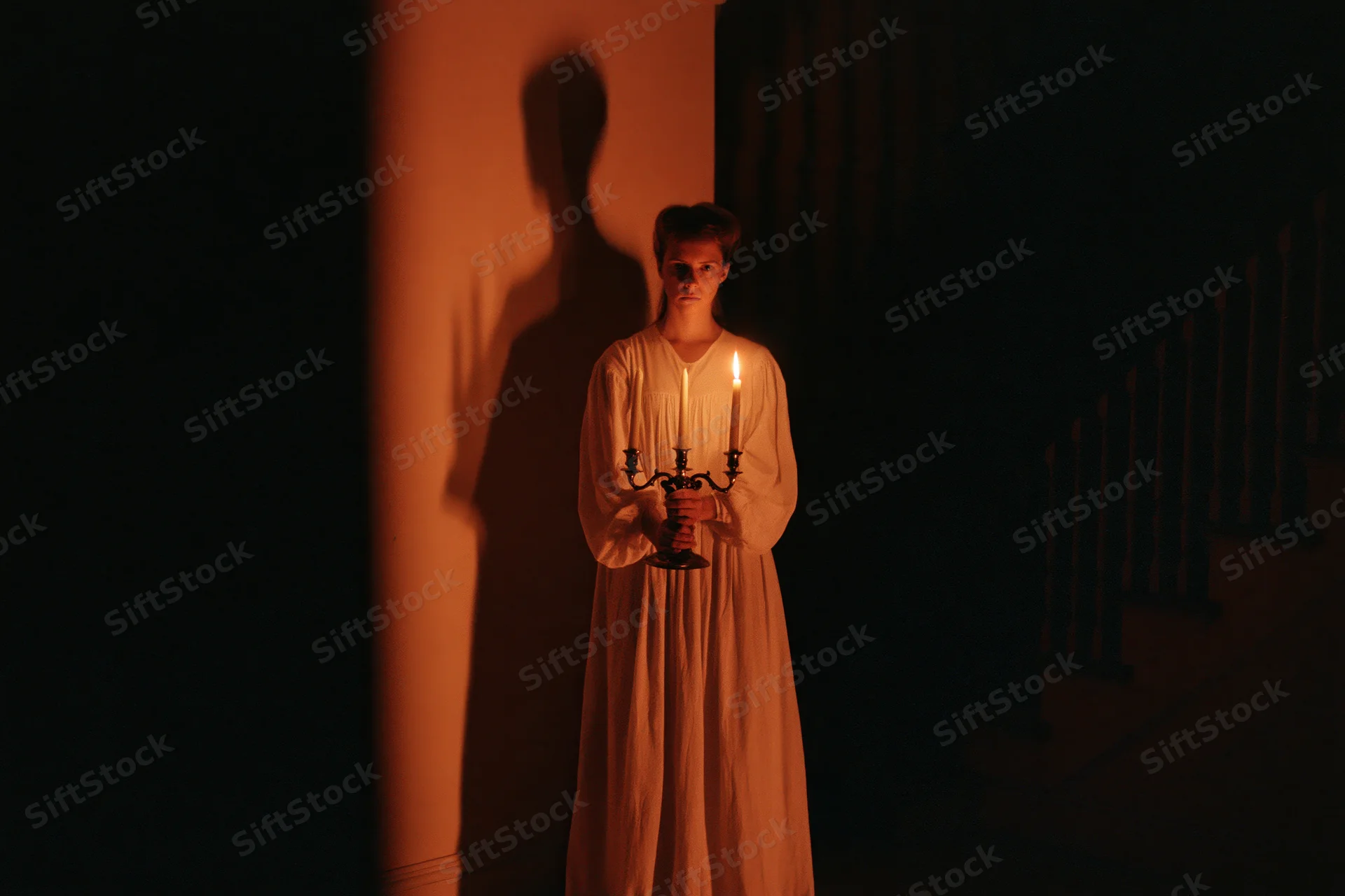 Woman in long white nightgown holding lit candle in dark hallway