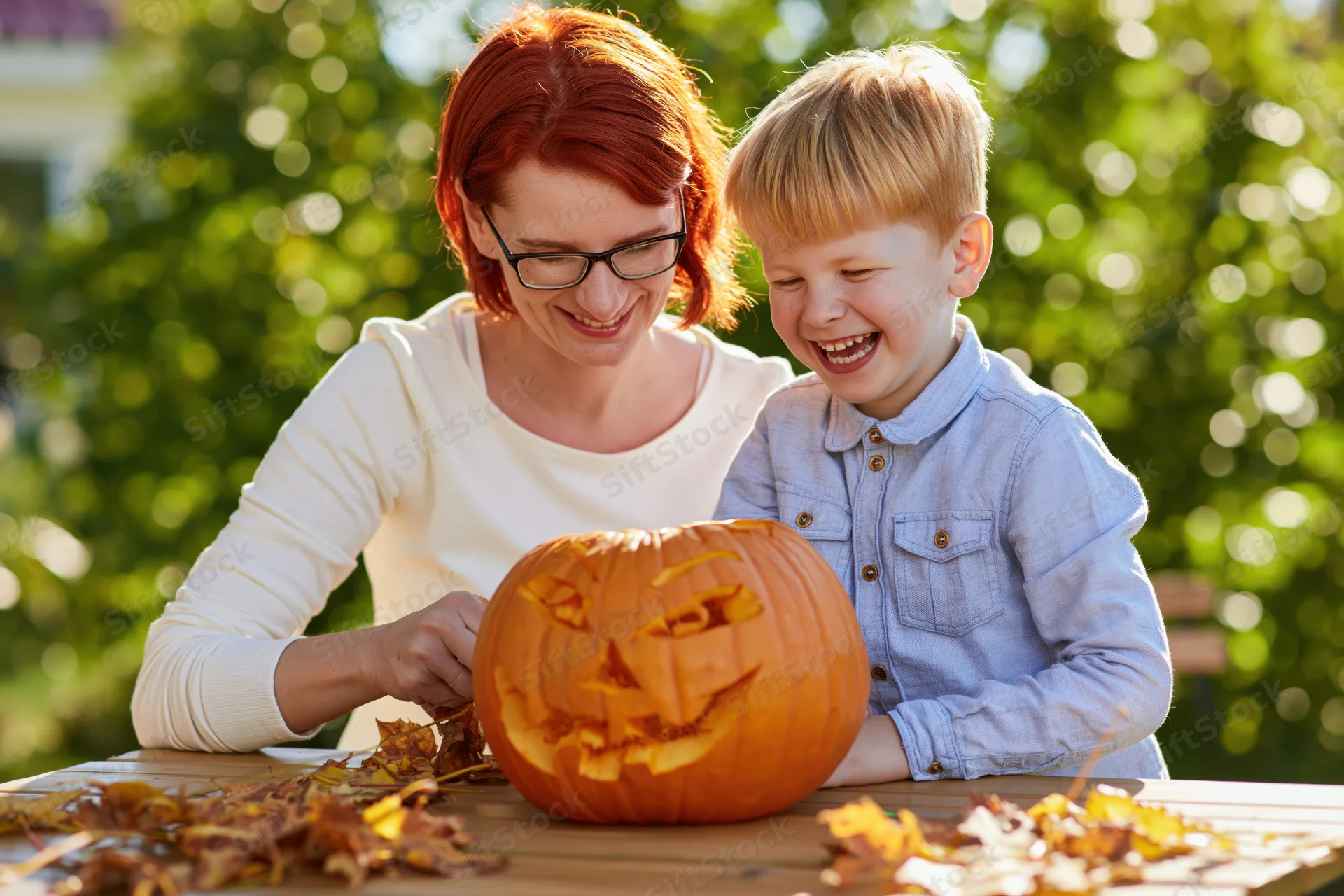 Woman and young boy carving a pumpkin together outdoors at a table surrounded by autumn leaves