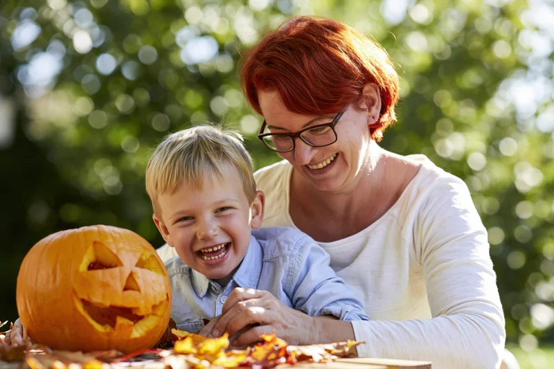 Woman and young boy smiling while carving a pumpkin outdoors in autumn foliage