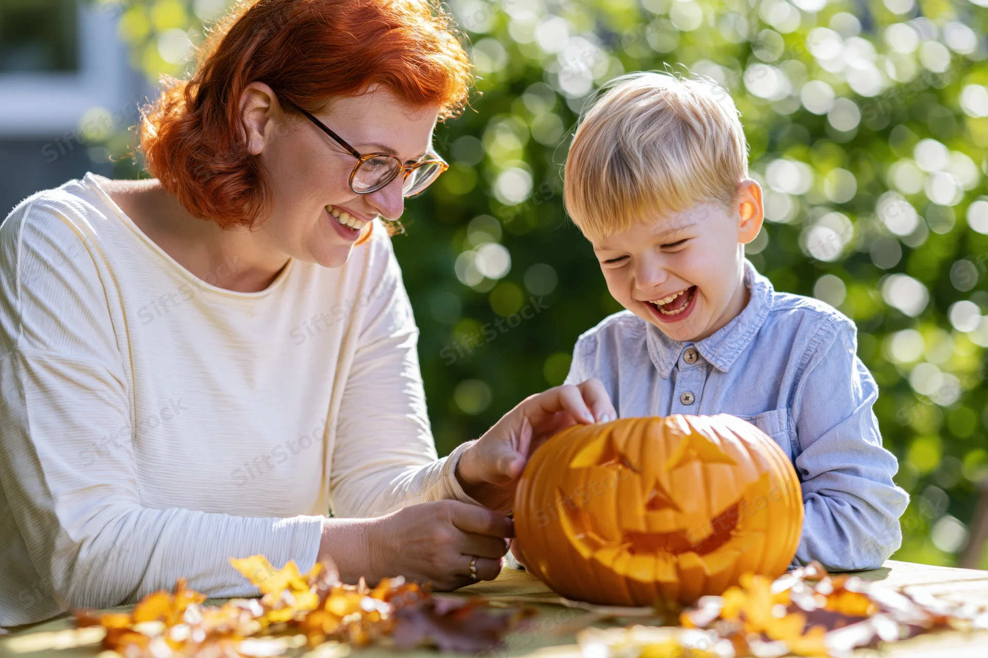 Woman and young boy laughing while carving a jack-o-lantern on a table outdoors surrounded by autumn leaves