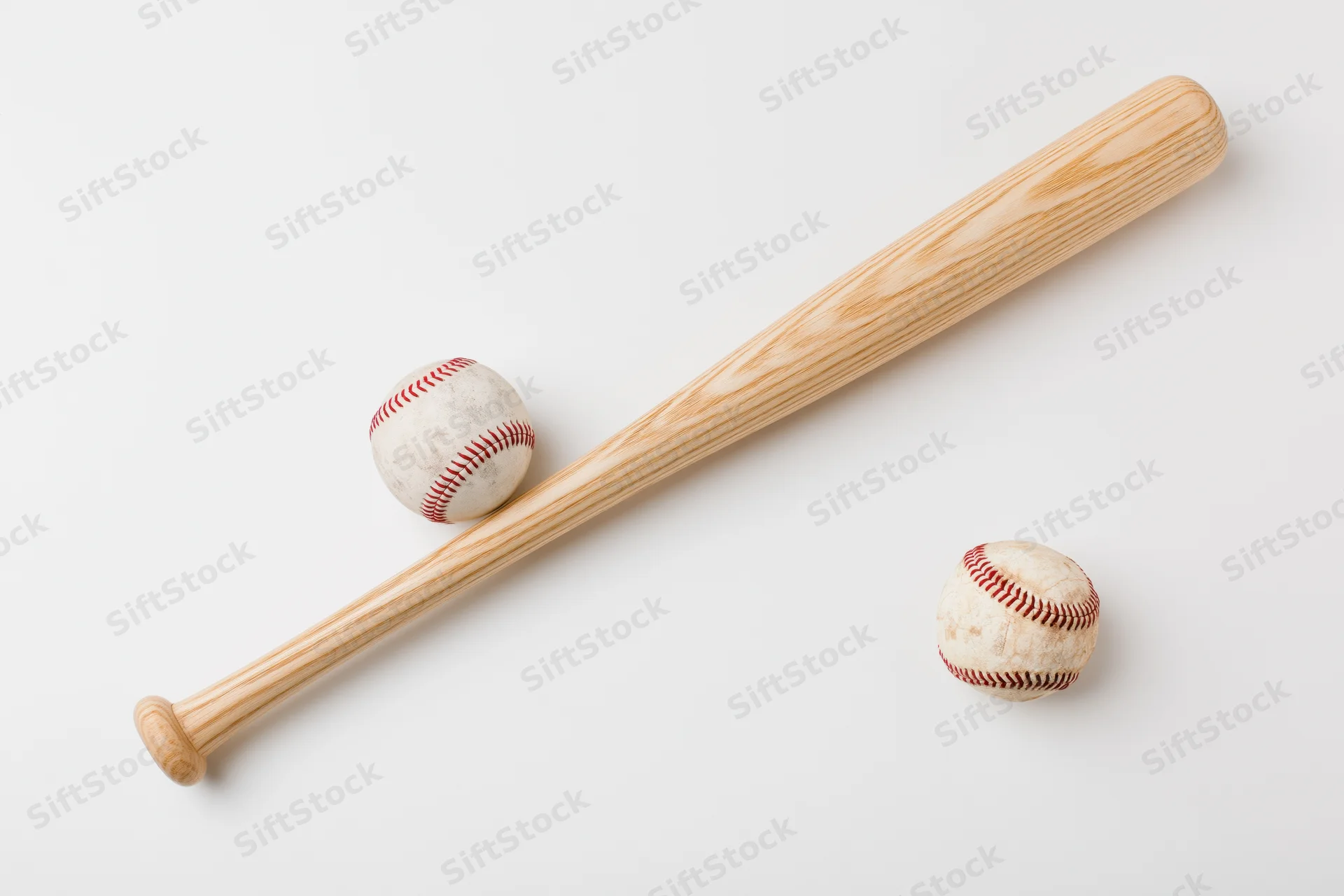 Wooden baseball bat lying diagonally beside two worn baseballs on white surface