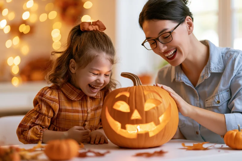 Woman and girl laughing while carving a glowing jack-o-lantern on a table indoors