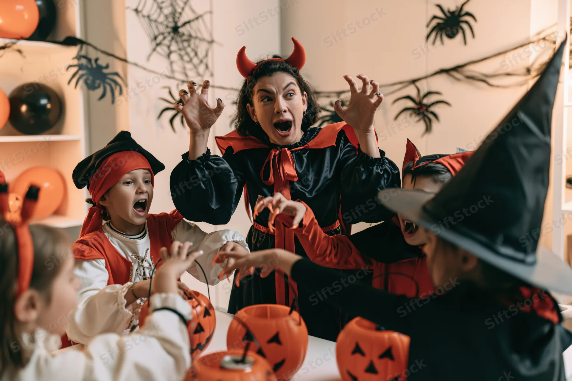 Woman in devil costume with children in halloween costumes at indoor party with pumpkins and decorations