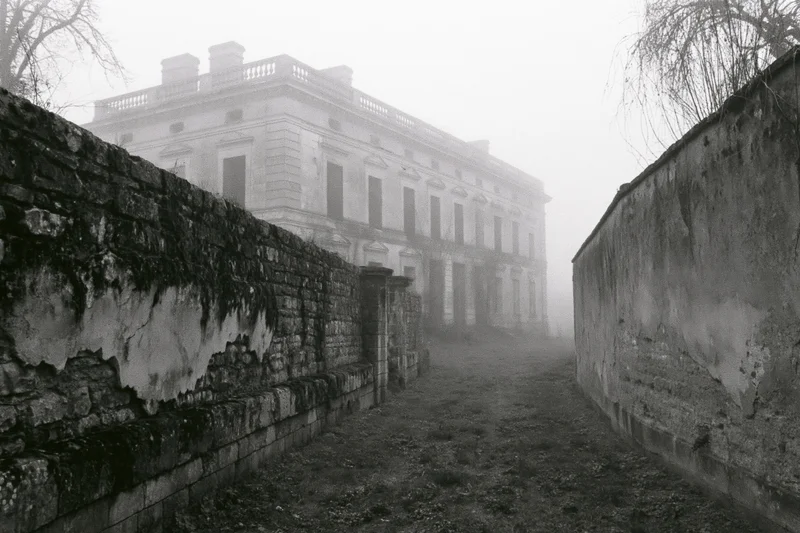 Abandoned neoclassical manor viewed through fog along a stone-walled pathway