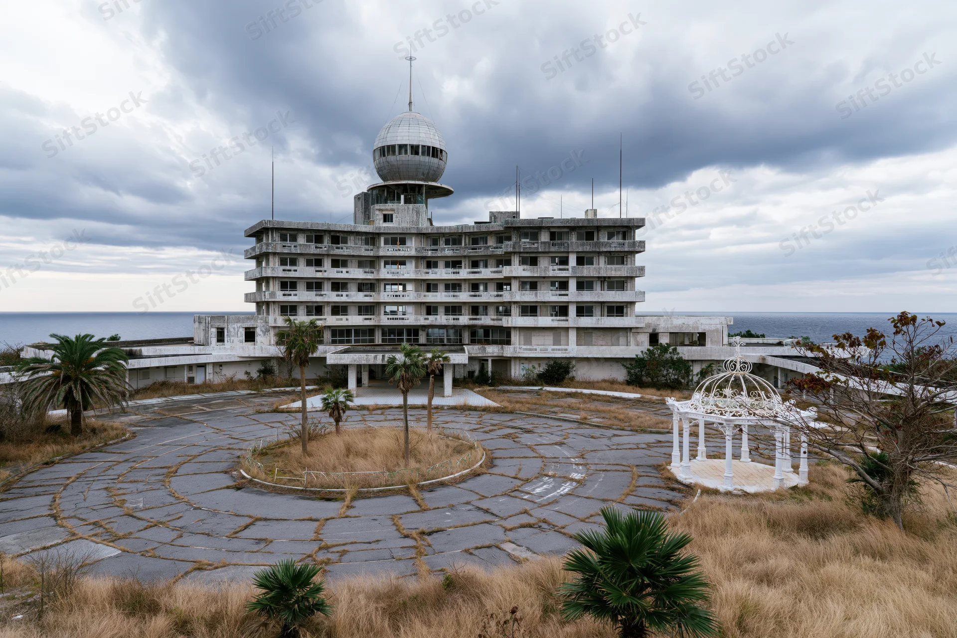 Abandoned japanese resort hotel with cracked circular driveway and white gazebo overlooking the ocean under stormy clouds