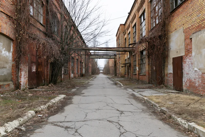 Narrow cracked asphalt pathway between abandoned brick industrial buildings with overgrown vines and metal overhangs