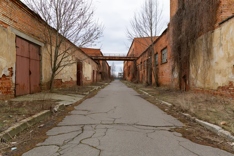 Cracked asphalt pathway between dilapidated brick buildings in an abandoned soviet-era industrial complex