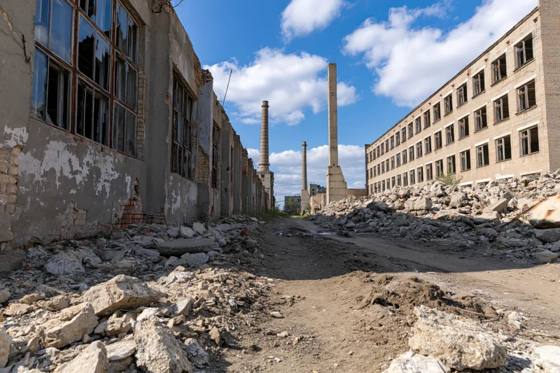 Wide alley between crumbling soviet-era factory buildings filled with rubble under a blue sky with clouds