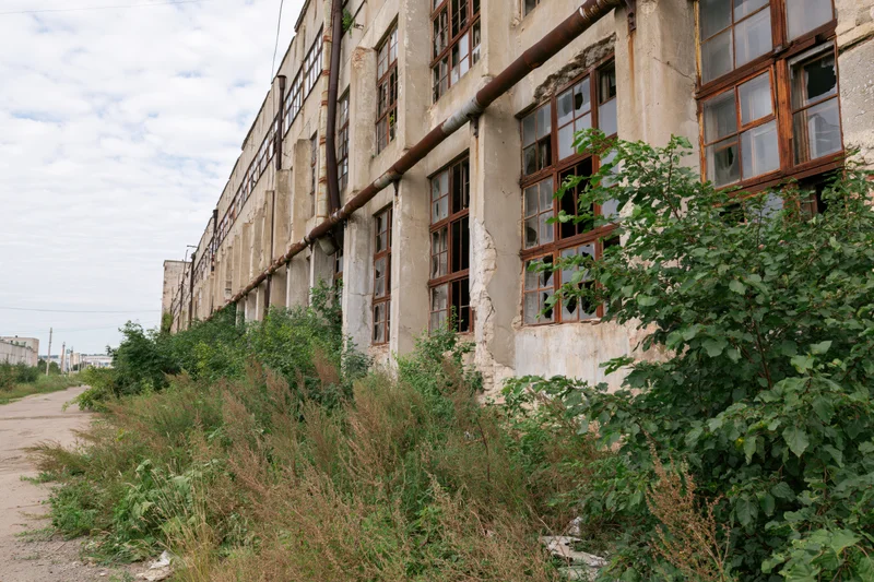 Long abandoned soviet-era industrial factory building with overgrown vegetation along its weathered facade