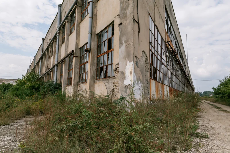 Abandoned soviet-era industrial factory building with broken windows and overgrown vegetation along a gravel path