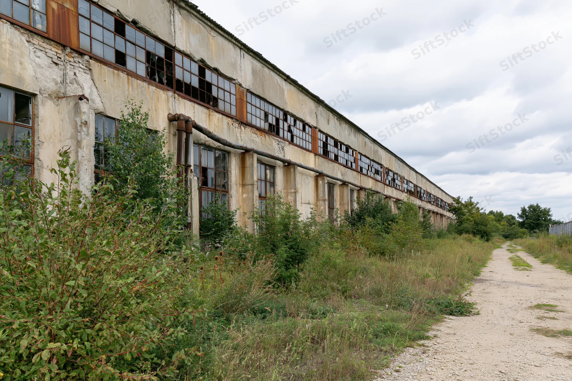 Long abandoned soviet-era industrial factory building with broken windows and overgrown vegetation along a dirt path
